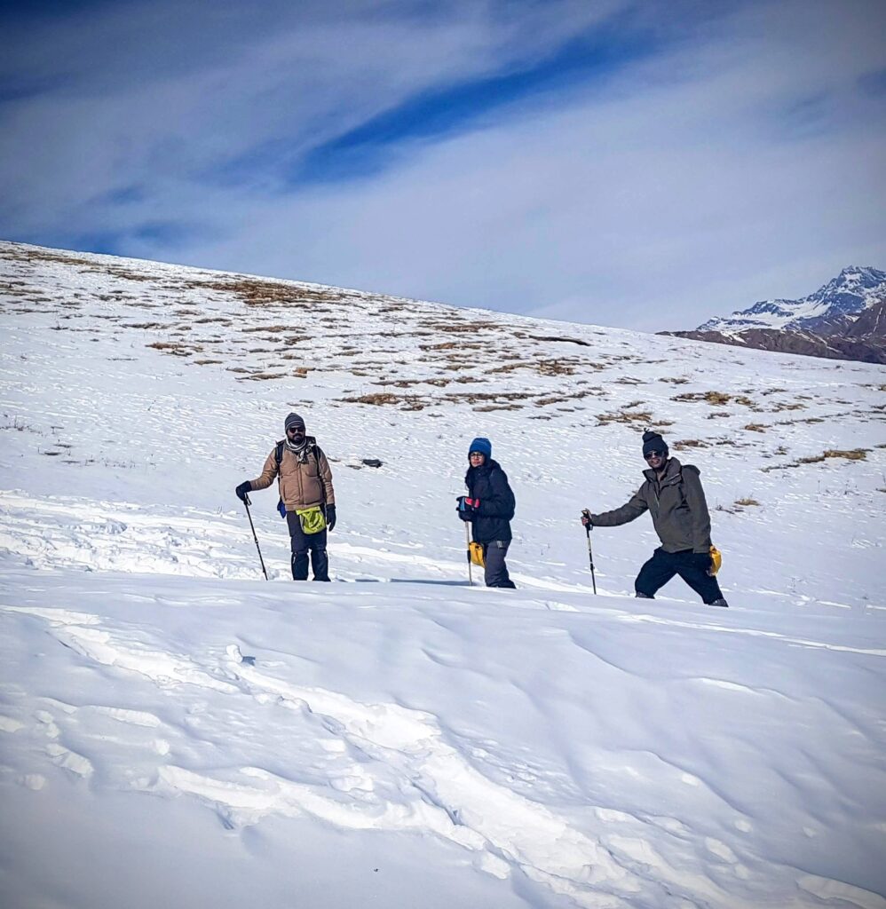 Three hikers, dressed in winter gear, trek across a snowy hillside under a partly cloudy sky. Snow-covered mountains are visible in the background. Each hiker carries trekking poles.