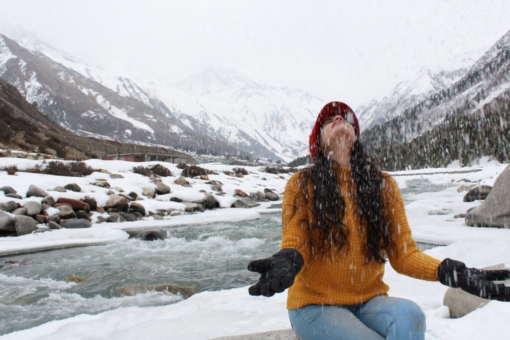 A person in a yellow sweater and red beanie joyfully tosses snow into the air, surrounded by a snowy mountain landscape and a flowing river. They are wearing black gloves, with the mountains and trees in the background dusted with snow.