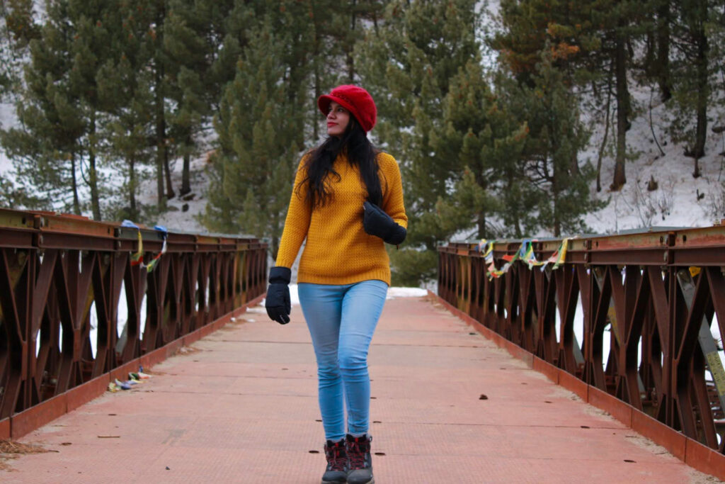 A person in a red hat and yellow sweater walks on a snow-dusted bridge surrounded by evergreen trees. Theyre wearing blue jeans, black gloves, and boots, enjoying a winter day in the scenic forest setting.